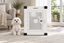 A Maltese sitting beside Impact's white Stationary Dog Crate in a living room on a woven rug.
