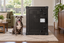A Pit Bull sitting on a woven rug beside Impact's black High Anxiety Crate in a living room.